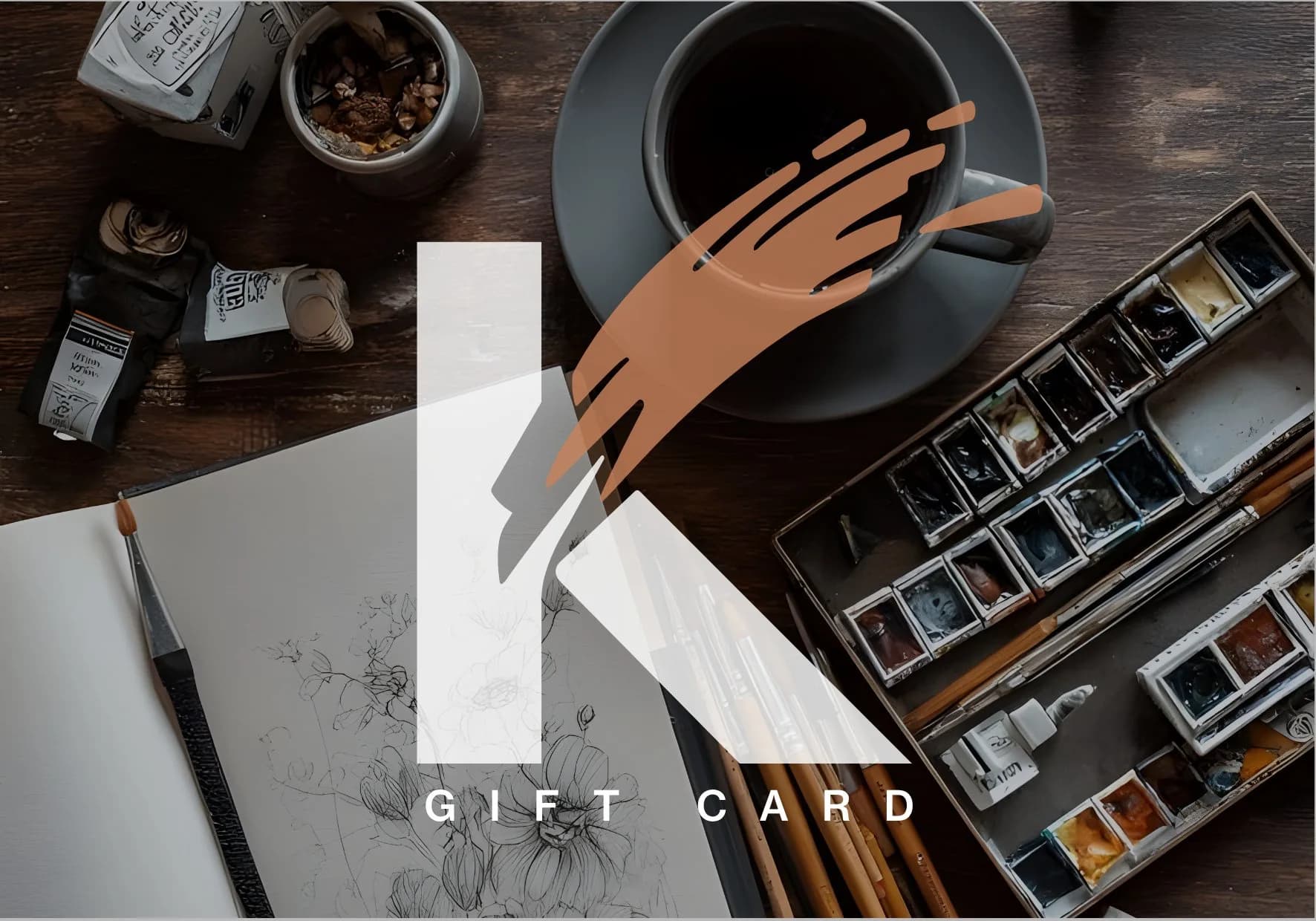 La Gift Card del Kreative Club: una grande K bianca con la pennellata terracotta su un tavolo con caffe', acquerelli e un album da disegno
