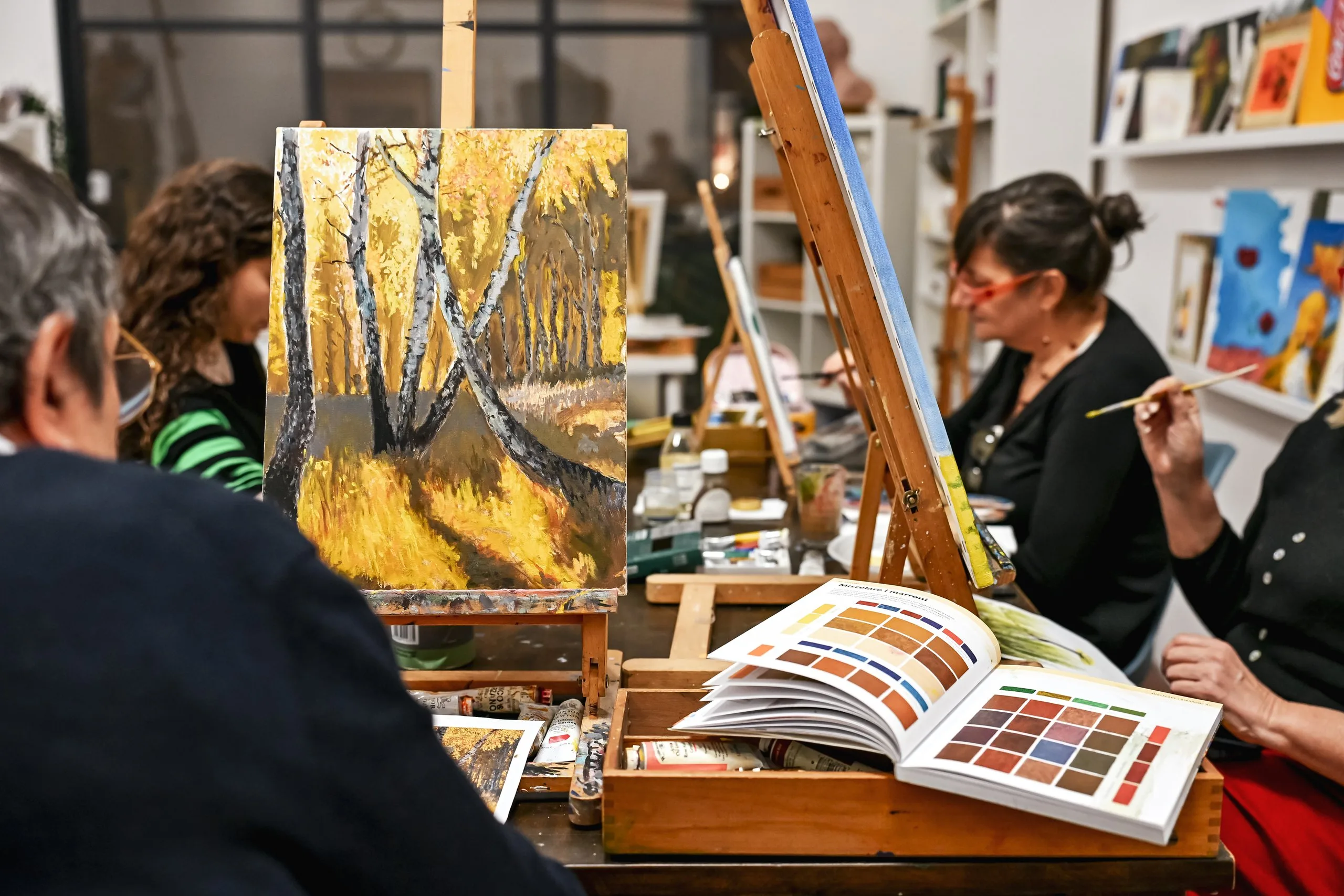 Allieve al cavalletto durante una lezione di pittura, con un libro di colori aperto sul tavolo