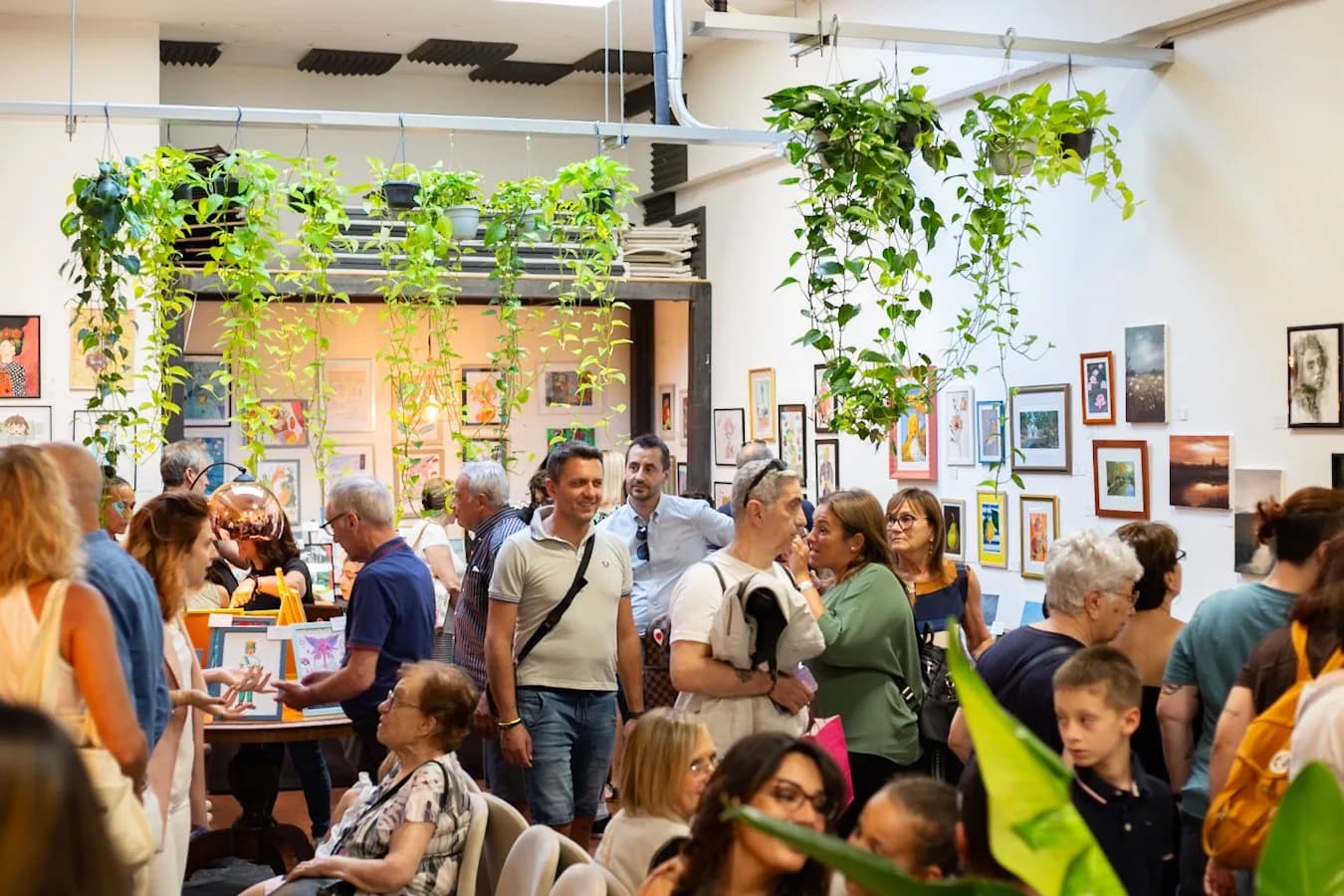 La sala della mostra piena di visitatori, con le piante appese sopra le opere