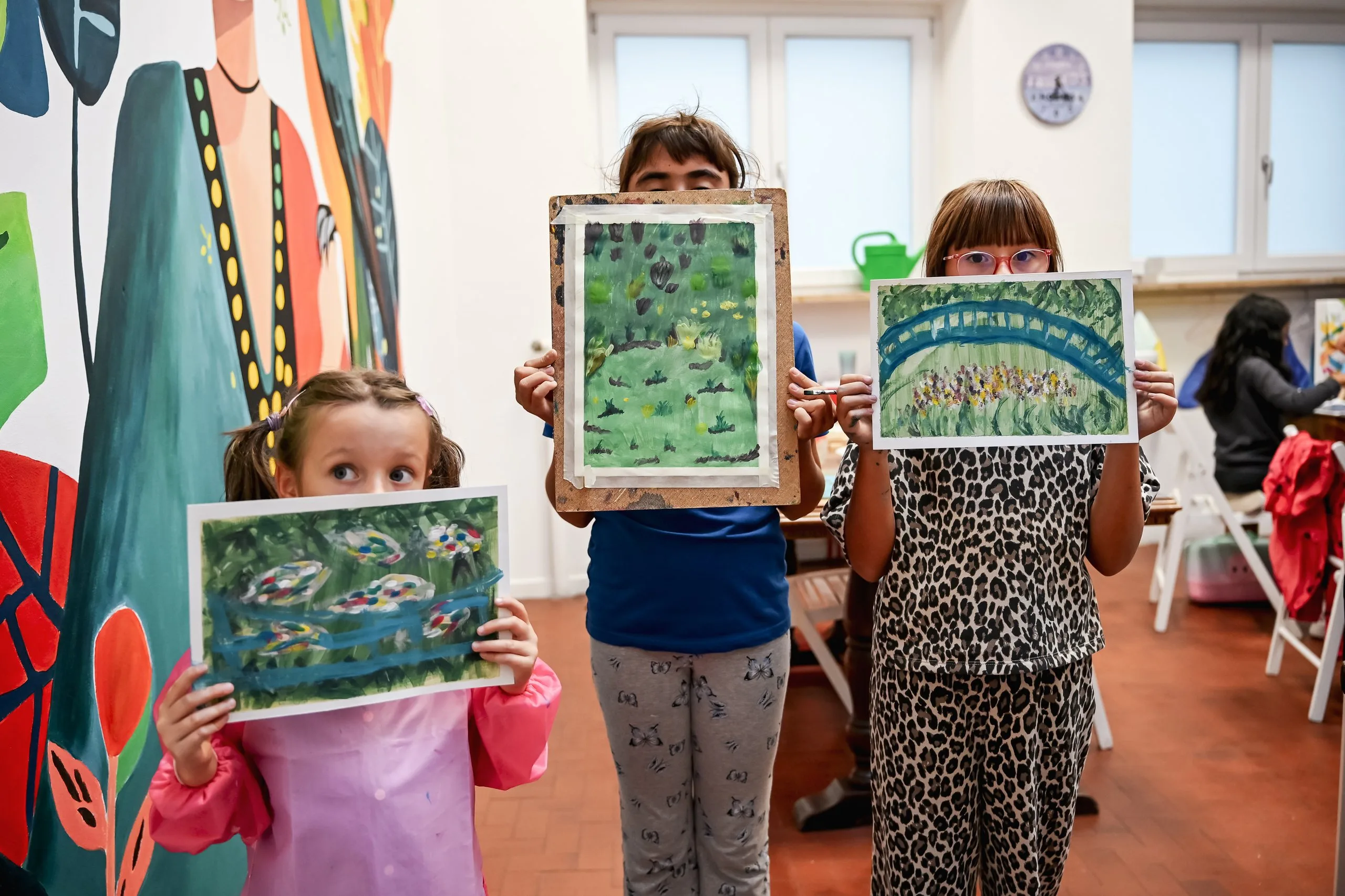 Tre bambine mostrano i loro dipinti di ninfee davanti a una parete dipinta